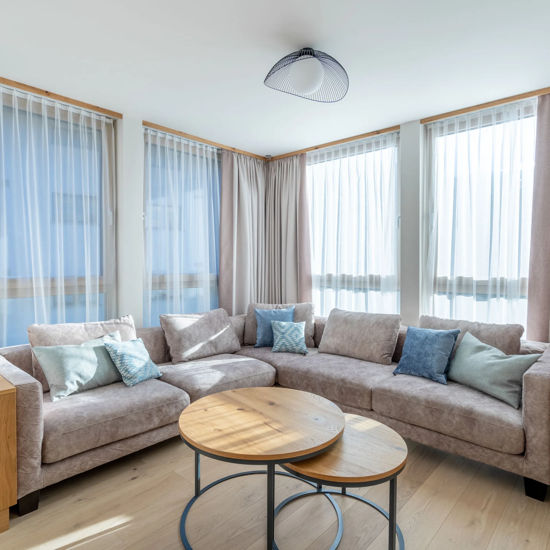 Helles Wohnzimmer mit grauem Ecksofa, Holz-Nestingtischen und großen Fenstern