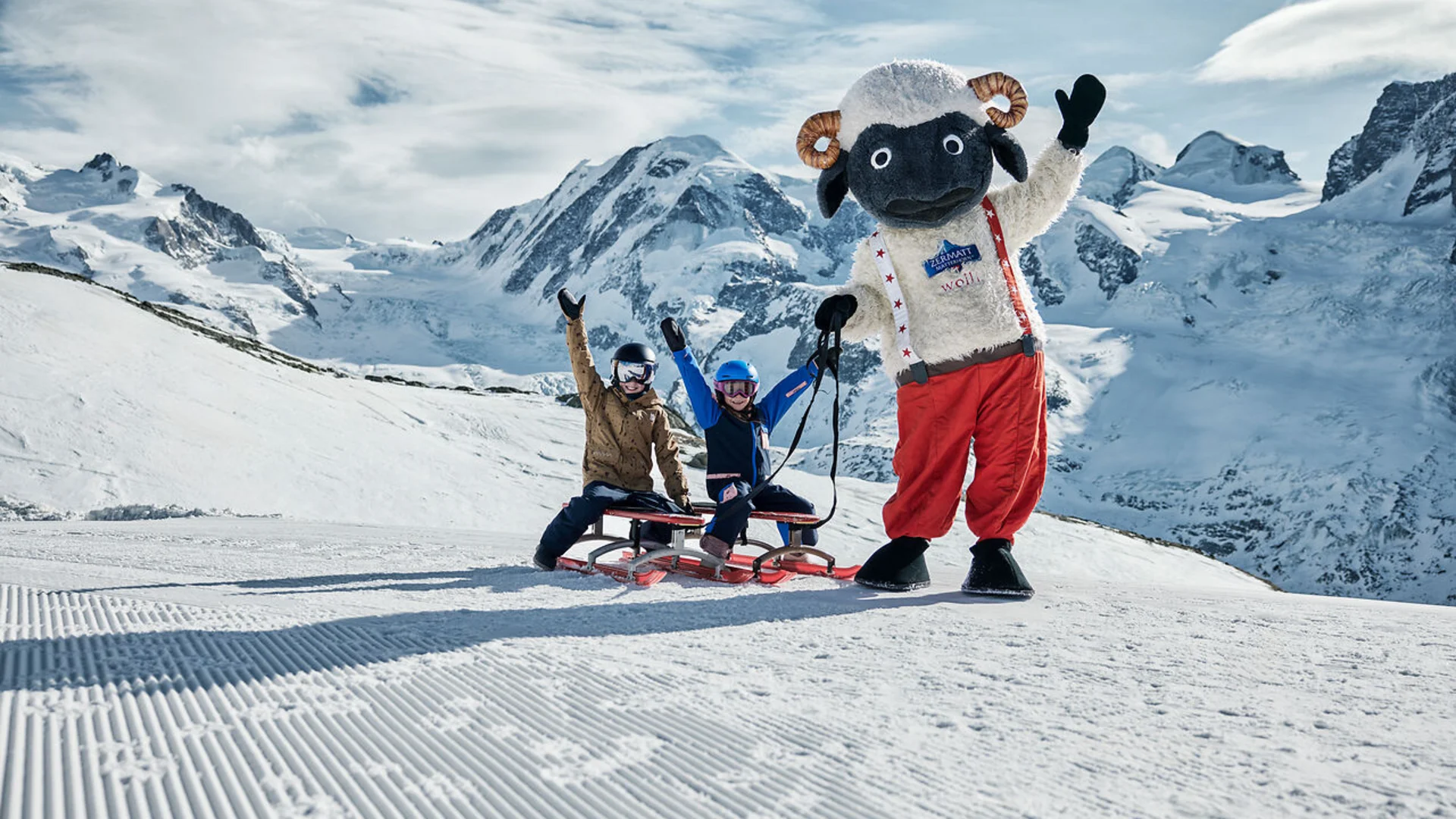 Two children on sled with standing sheep mascot in snowy mountain landscape