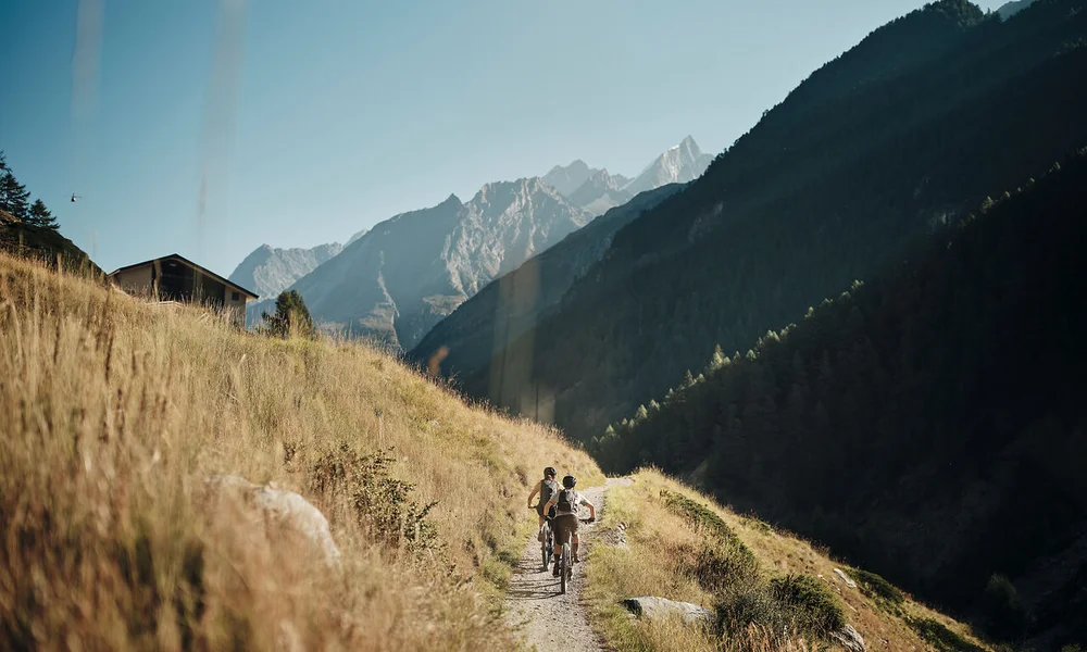 Zwei Radfahrer fahren auf einem Bergweg mit Alpen im Hintergrund