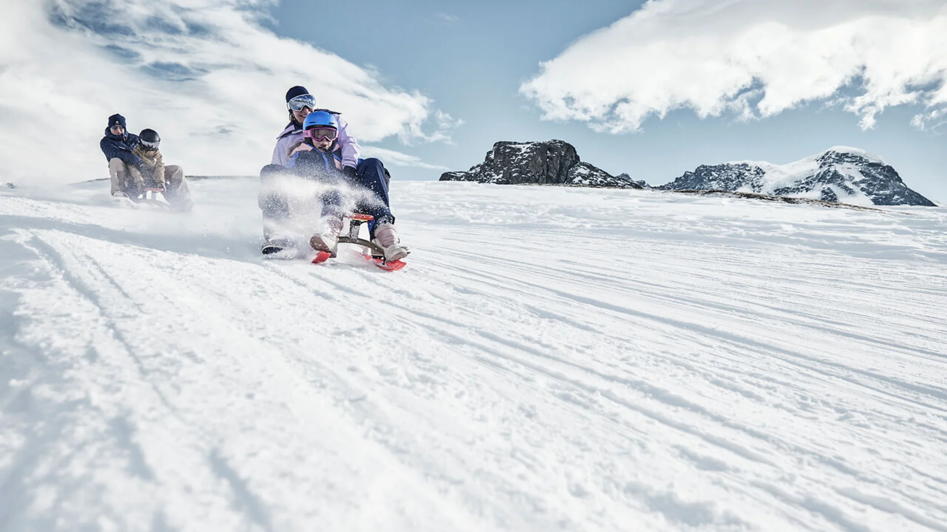 Four people sledding down a snowy hill with mountains in the background