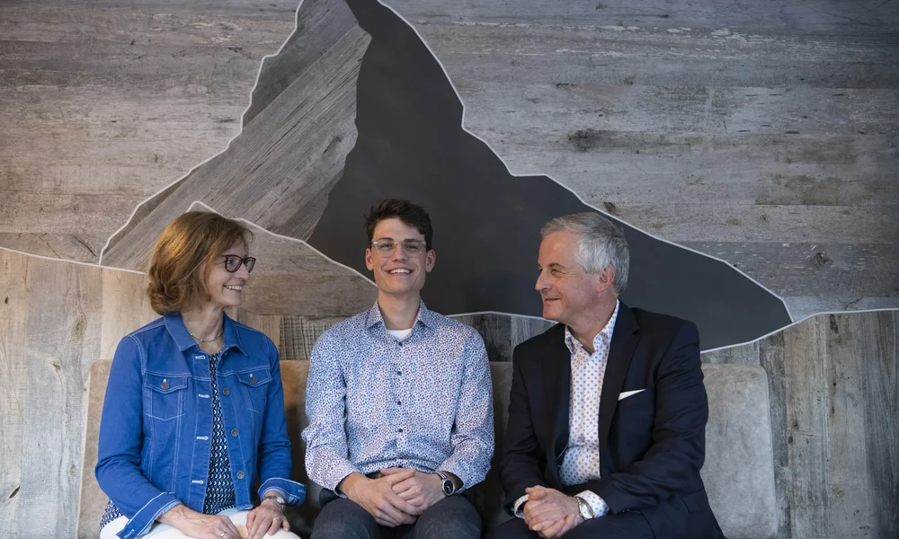 Rooms, Apartments & Suites - Resort La Ginabelle Zermatt Seated host family in front of a wooden wall cladding with Matterhorn design