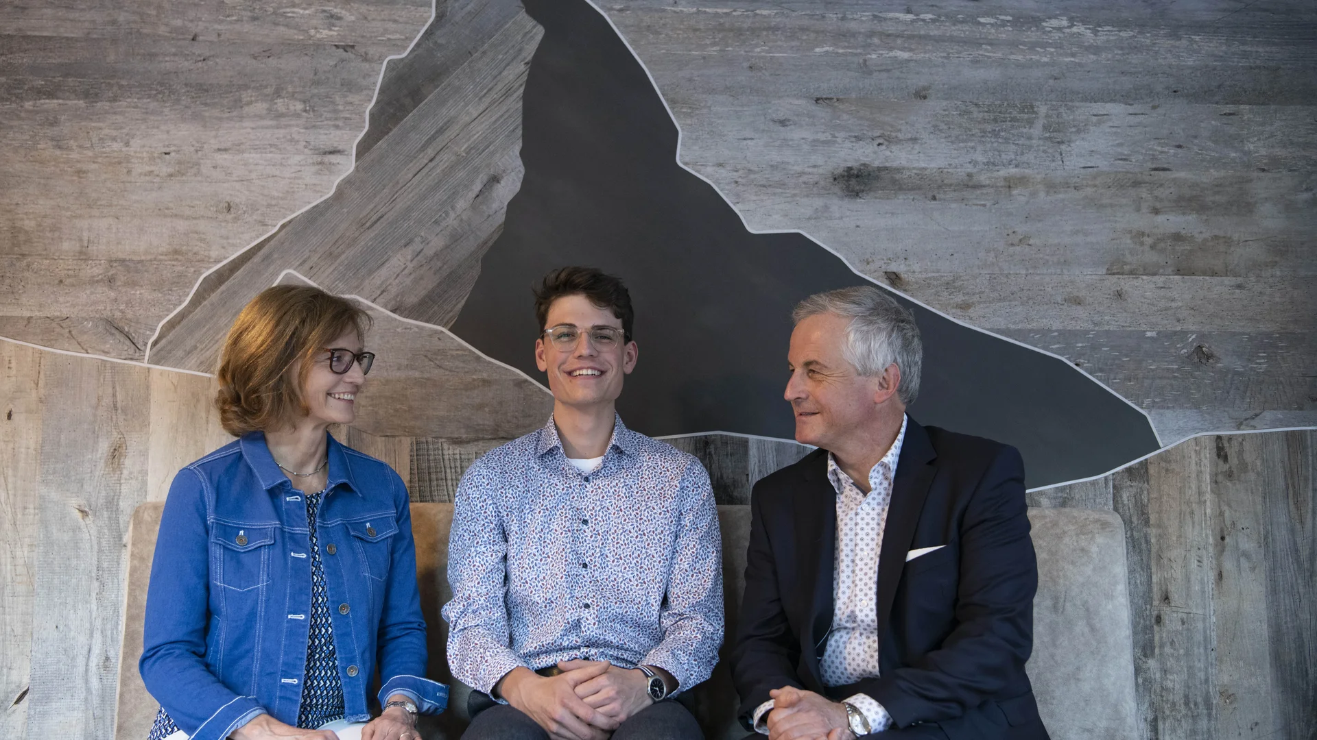 Sitzende Gastgeberfamilie vor einer hölzernen Wandverkleidung in Matterhornoptik