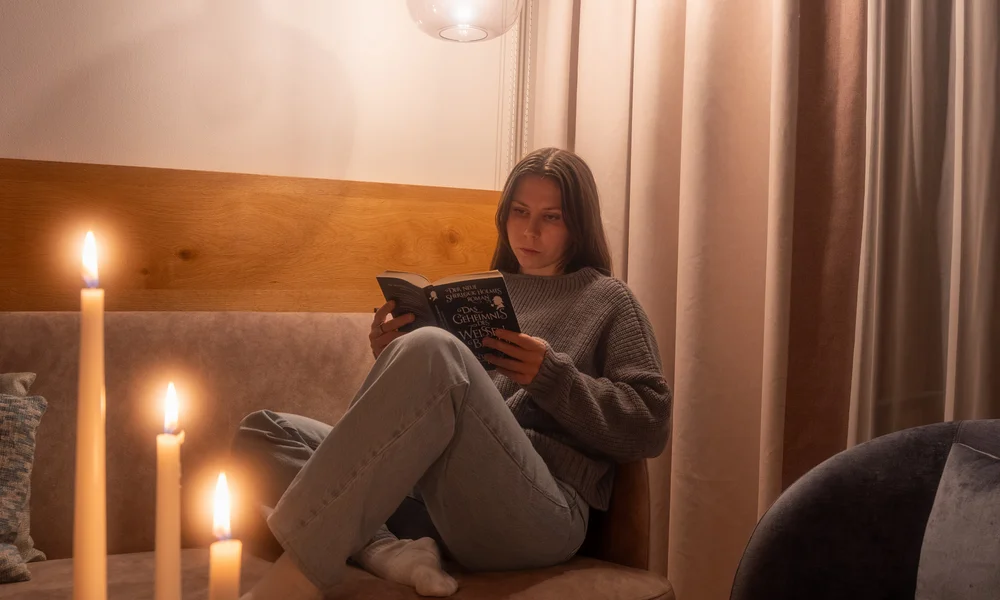 acotar Frau liest Buch auf Sofa bei Kerzenlicht und warmem Lampenlicht
