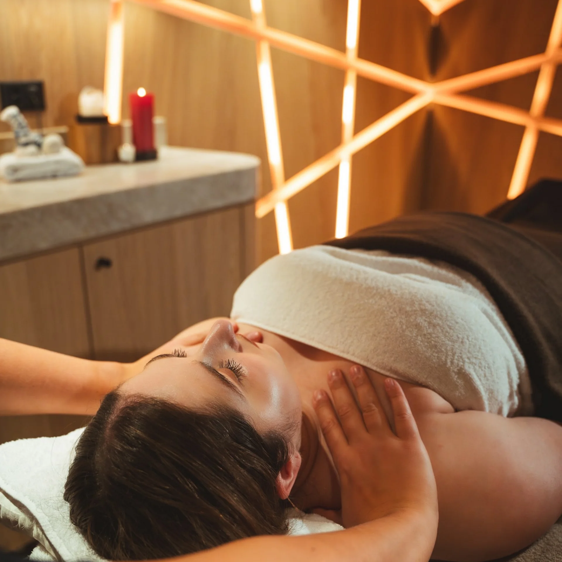 Nackenmassage bei einer Frau in einem Behandlungsraum mit beleuchteter Wandverkleidung.