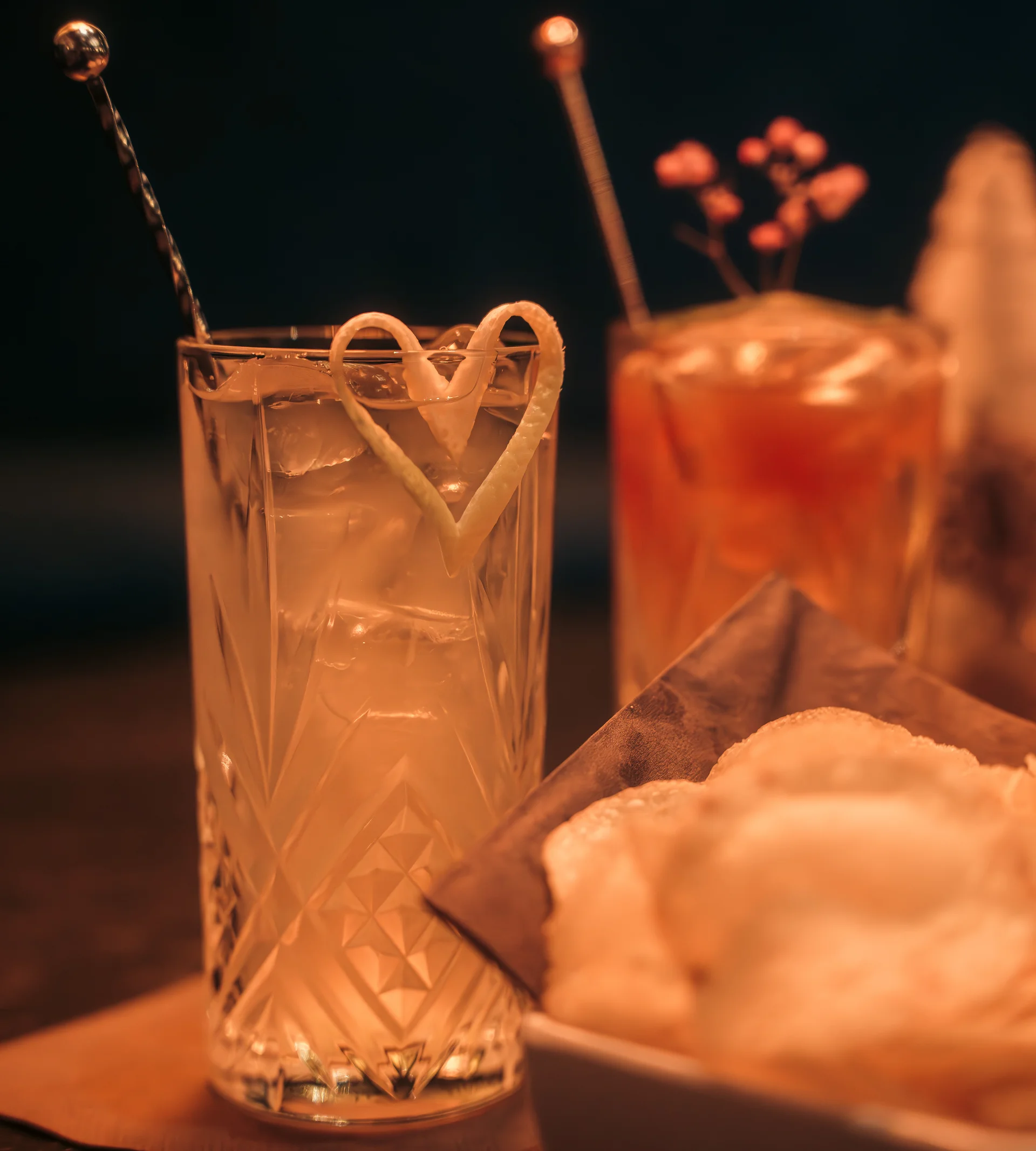 acotar Cocktails mit Eis und Zitronenschale, daneben Teller mit Chips bei gedämpftem Licht