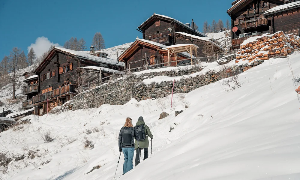 Wintersport Zwei Wanderer steigen in winterlicher Berglandschaft zu verschneiten Holzchalets auf