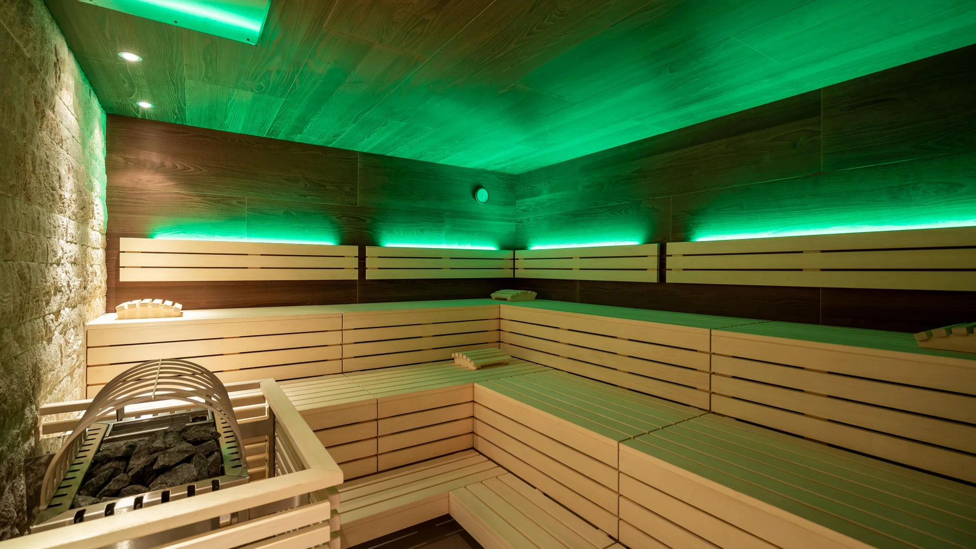 Wellnessresort in Zermatt - Resort La Ginabelle Zermatt Bio Sauna mit indirekter grüner Beleuchtung, Holzbänken und in Holz verkleiderter Ofen.