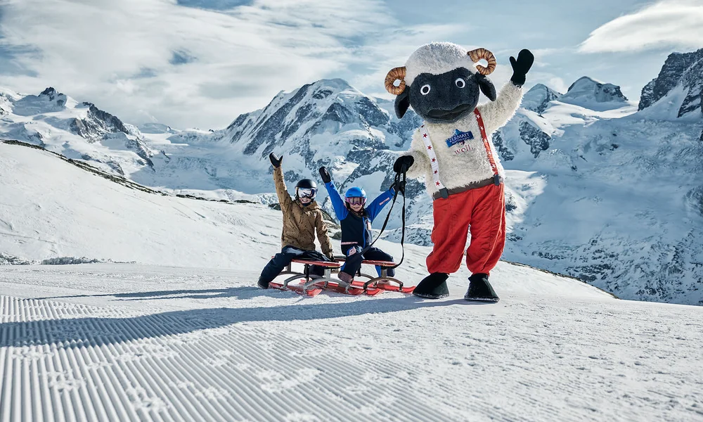 Deux enfants sur des luges avec une mascotte de mouton dans un paysage de montagne enneigé