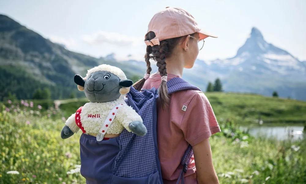 Enfant avec casquette rose et sac à dos avec peluche Wolli devant paysage montagneux